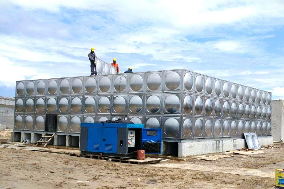 Workers standing on a large stainless steel modular water tank installed outdoors with equipment nearby