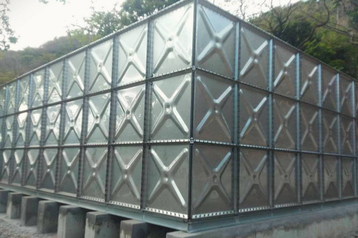 Large stainless steel modular water storage tank installed outdoors on concrete supports near a hillside